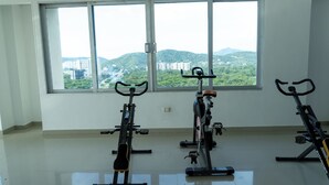 Une salle équipée de trois vélos d’appartement, de grandes fenêtres donnant sur un paysage verdoyant et des bâtiments, et dotée d’un sol carrelé.