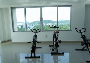 A room with three stationary bikes, large windows with a view of greenery and buildings, and a tiled floor. - Modern apartment with pool in Bello Horizonte | Near the beach (Santa Marta)