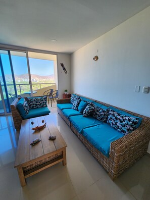 A living room with a wicker sofa, a wooden coffee table, and a view of a balcony. - Modern apartment with pool in Bello Horizonte | Near the beach (Santa Marta)