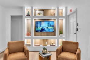 A modern living room with a flat-screen TV mounted on the wall, two armchairs, a coffee table with books, and decorative items on shelves. - Stylish 2 br, bbq, Close to beach, Bal Harbor, Aventura Mall, Design District. (Biscayne Park)