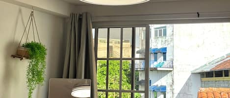 A room with a ceiling fan, a hanging plant, a window with a view of buildings, and a light fixture.