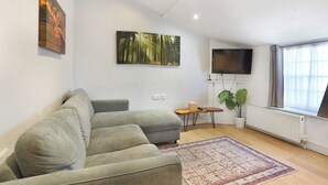 A modern living room with a grey sectional sofa, a large forest-themed painting, a flat-screen TV, a small wooden side table, and a potted plant. - Central London 1-Bed Flat – Hyde Park & Paddington Area (London)