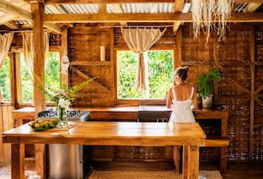 A rustic wooden interior with a large wooden table, a kitchen area, and a view of greenery outside.