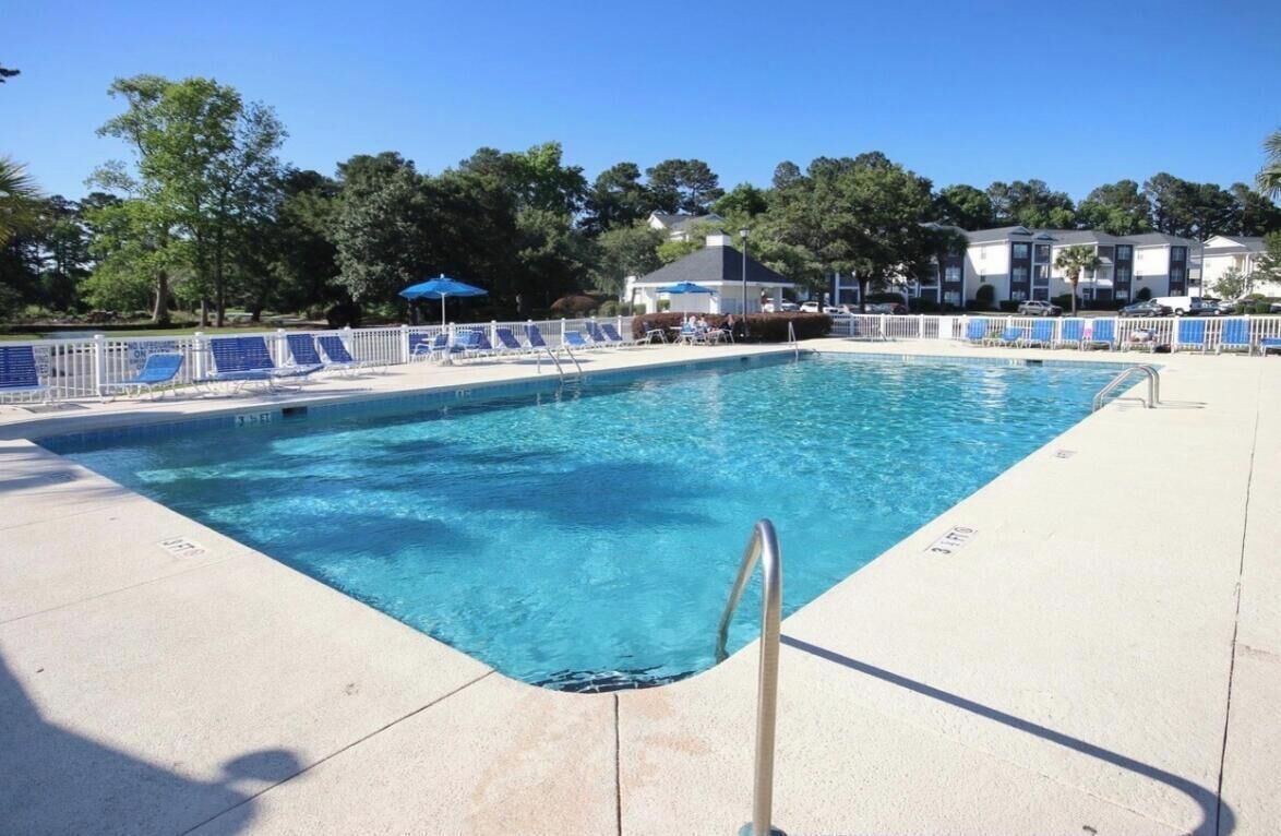 A rectangular swimming pool with a metal ladder, surrounded by a white fence and blue lounge chairs.