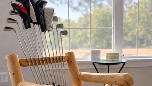 Una silla de madera con palos de golf apoyados, ubicada en una habitación con un ventanal grande y una mesita.