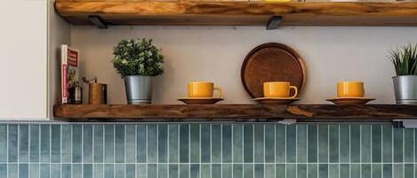 Una cocina con salpicadero de azulejos azules, repisa de madera con plantas en macetas, tazas amarillas y una cafetera.