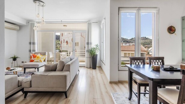 Uma sala ampla com sofá, mesa de jantar e cadeiras, janelas grandes com vista para prédios e colinas, e uma planta em vaso.