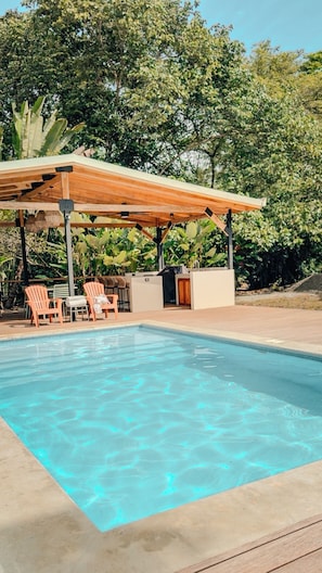 Piscina com pergolado de madeira, cadeiras de descanso e um bar.