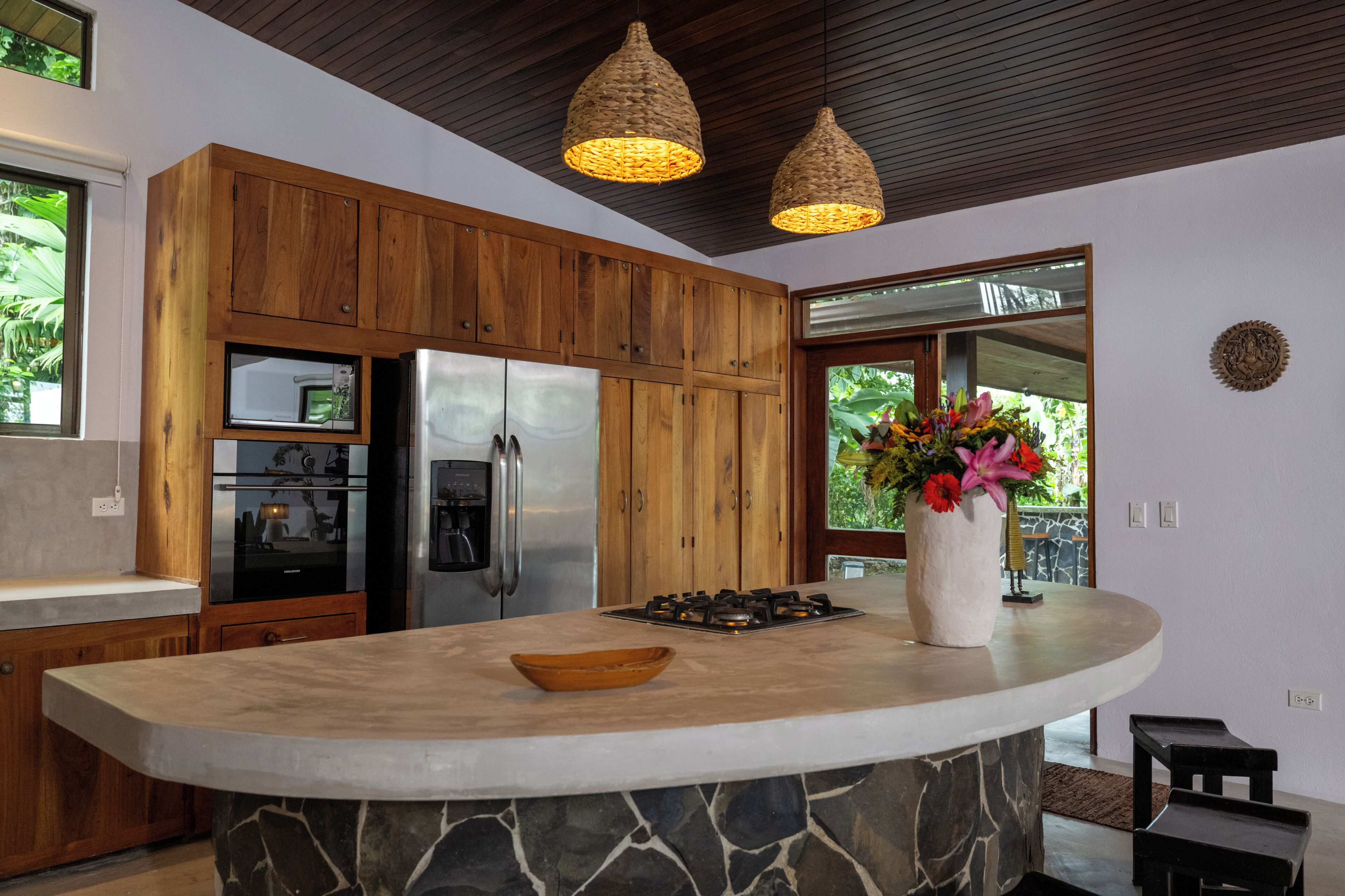 Una cocina con armarios de madera, una isla de piedra, un refrigerador, un microondas y un florero con flores.