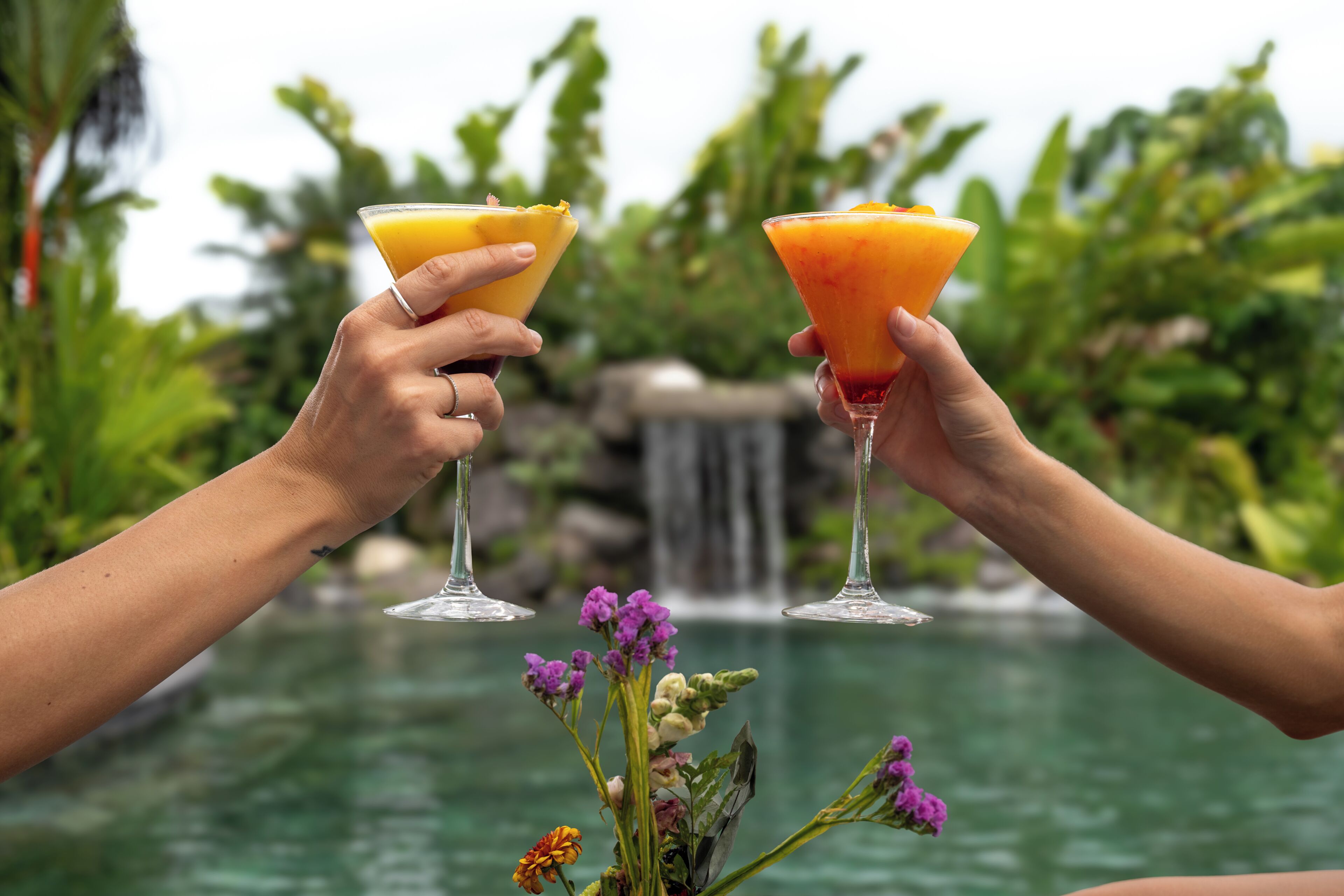 Two hands holding cocktail glasses with tropical drinks, a waterfall, and lush greenery in the background.