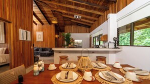 Uma sala de jantar com vigas de madeira, uma cozinha ao fundo e vista para o verde pela janela.