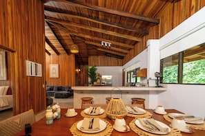 Comedor con vigas de madera, cocina al fondo y vista a la vegetación a través de una ventana.