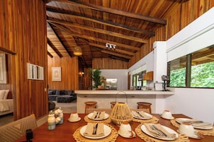 Uma sala de jantar com vigas de madeira, uma cozinha ao fundo e vista para o verde pela janela.