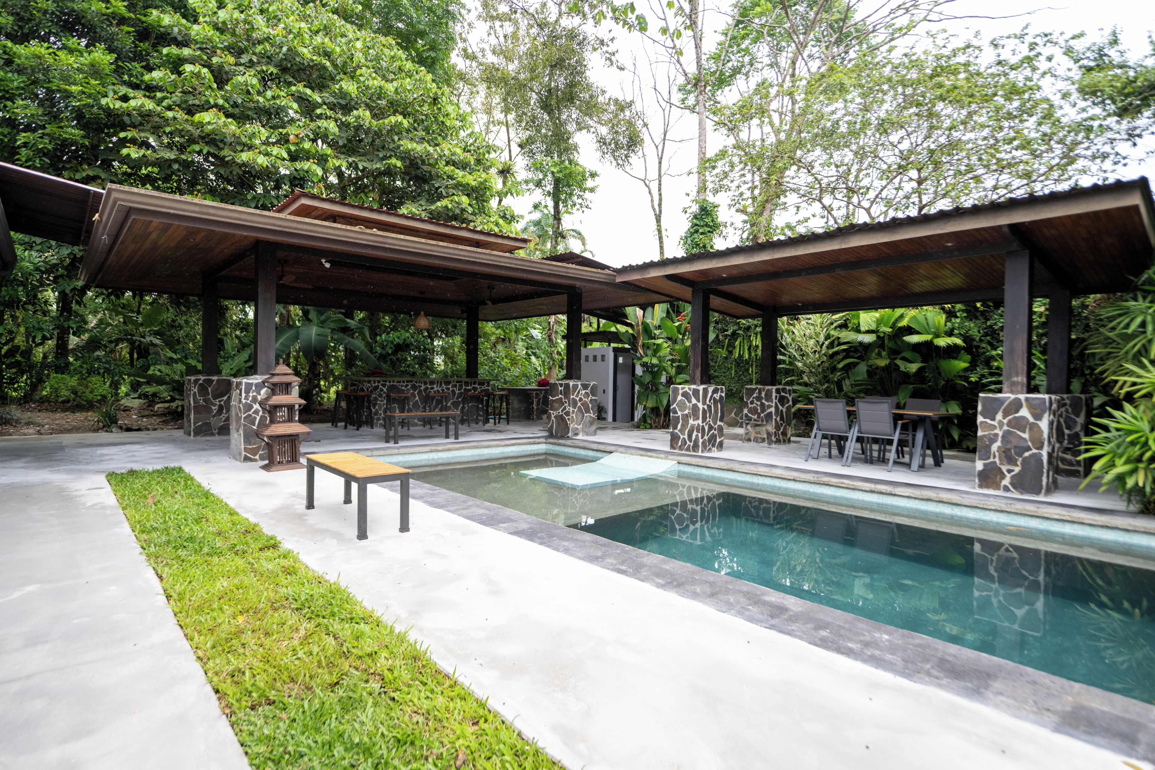 Un área de piscina con un porche cubierto, muros de piedra y mobiliario exterior.
