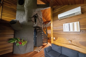Interior de una casa en árbol con techo de madera, un sofá gris y un aire acondicionado fijado a la pared.