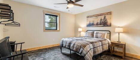 A bedroom with a bed, a desk, a chair, a ceiling fan, a window, and a painting on the wall.