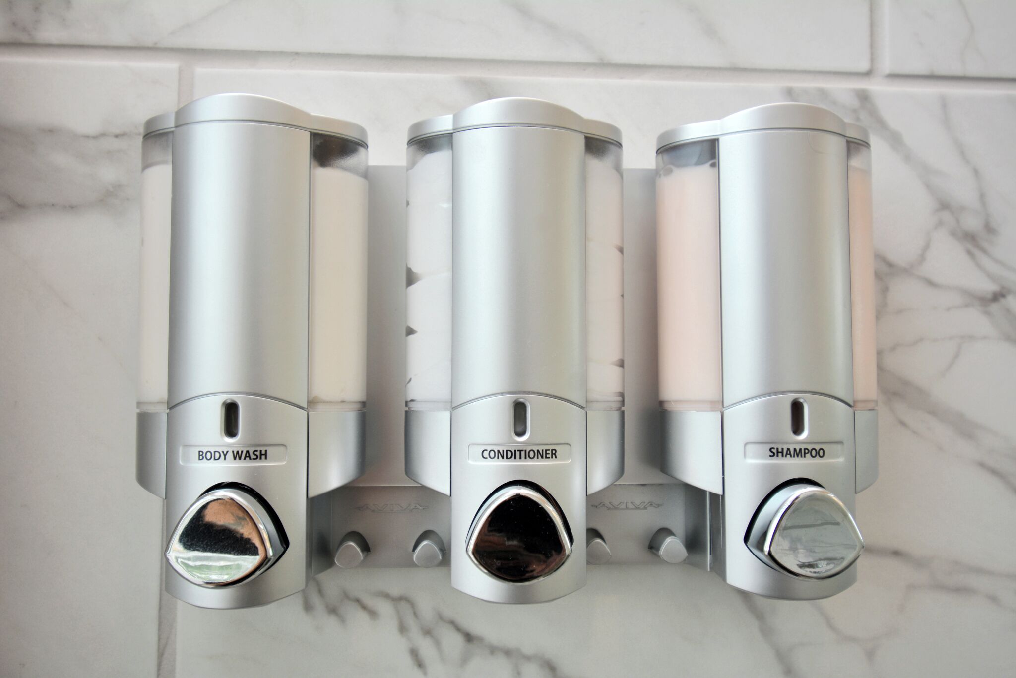 Three metallic dispensers labeled BODY WASH, CONDITIONER, and SHAMPOO in a bathroom.