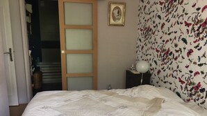 Ein Schlafzimmer mit einem Bett, einem Nachttisch, einer Lampe, einem gerahmten Bild an der Wand und einer Tür zu einem anderen Raum.