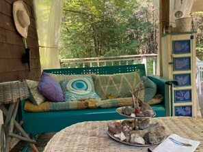 Un área de estar al aire libre acogedora, con un sofá verde, almohadones decorativos, una mesa de mimbre con rocas y un gato, y una pared de madera con un sombrero colgado.