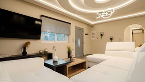 Een moderne woonkamer met een flatscreen televisie, witte leren banken, een houten salontafel en een plafond met ronde verlichting.