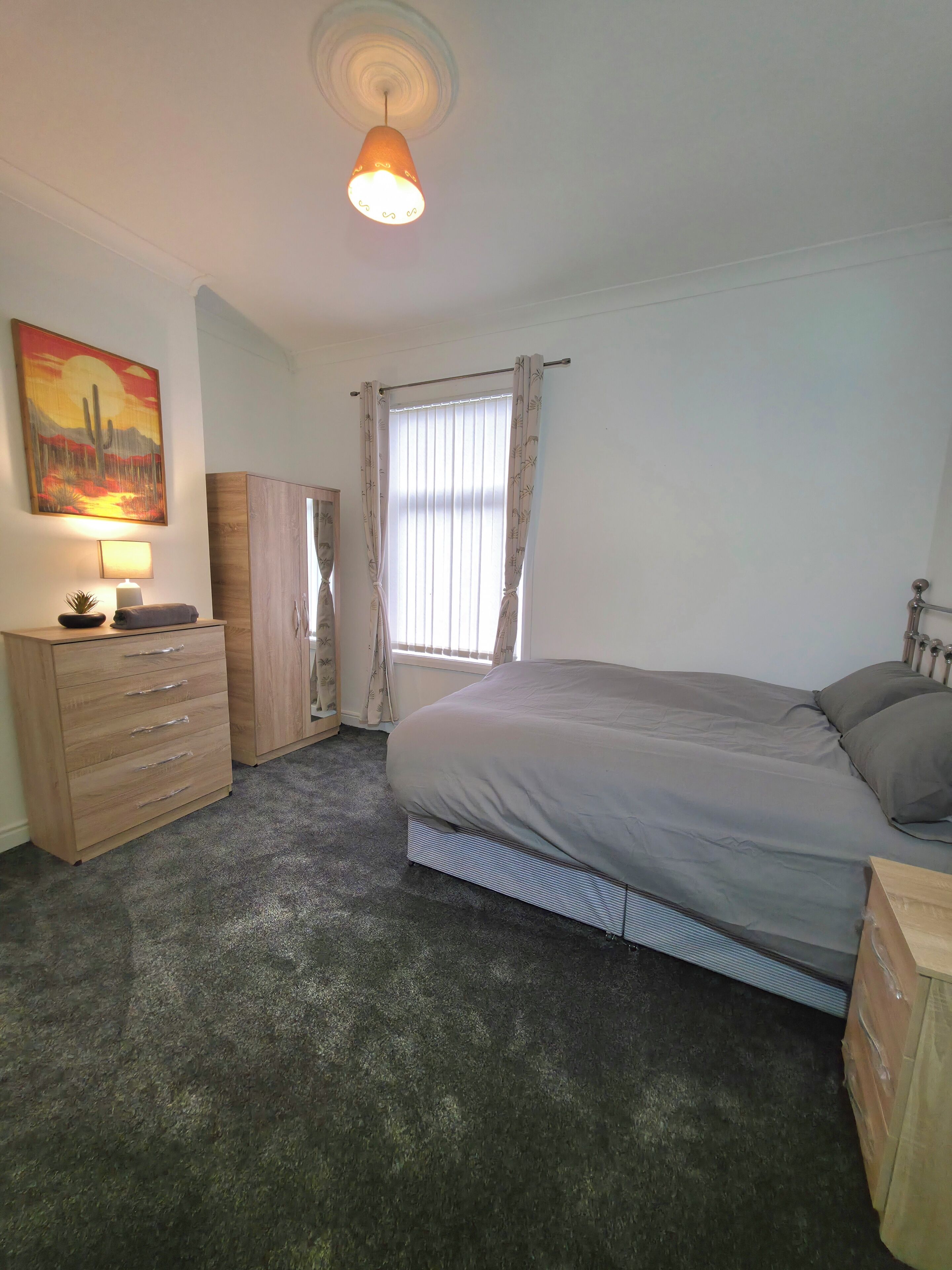 A bedroom with a bed, a dresser, a nightstand, a lamp, a window with curtains, and a painting on the wall.