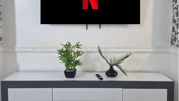 A flat-screen TV mounted on a wall with a large red 'N' on the screen, a white console table with two potted plants, and a remote control on the table. - Costa Home Middlesbrough (Middlesbrough)