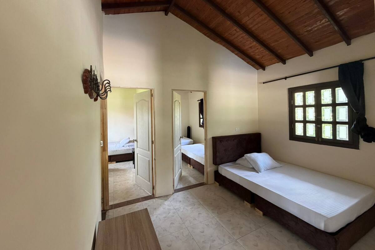 A room with a wooden ceiling, a bed with a headboard, a window with curtains, and a door leading to another room.