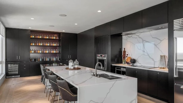 Una cocina moderna con una isla de mármol, taburetes de bar, una vinoteca empotrada y un refrigerador de acero inoxidable.