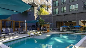 Una piscina con sillas de descanso y sombrillas, rodeada por un edificio moderno con balcones.