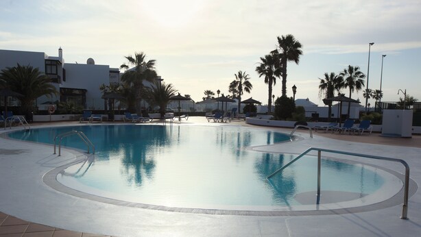 En swimmingpool med ett kaklat poolområde, solstolar, palmer och en byggnad i bakgrunden.