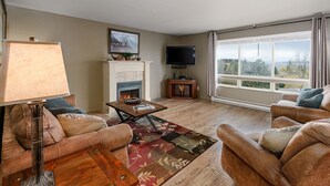 A living room with a fireplace, a television, a sofa, a coffee table, and a large window with a view. - Marina Cay Condo Retreat (Bigfork)