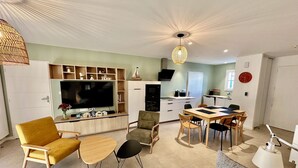 Una sala moderna con televisor de pantalla plana, muebles de madera, un comedor con mesa y sillas, y una cocina al fondo.