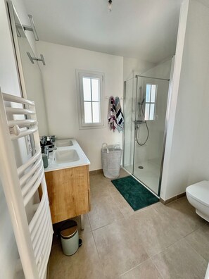 Un baño moderno con cabina de ducha de vidrio, tocador de madera, lavamanos blanco y un pequeño cubo de basura.