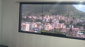 Une chambre avec une grande fenêtre, un lit recouvert d’une courtepointe à motifs et une vue sur un paysage urbain avec des bâtiments et une montagne.
