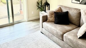 A living room with a beige sofa, a flat-screen TV mounted on the wall, a potted plant, and a large window with a view of the outside. - FIFA Fans! Brand-NEW 1 Bedroom, 1 Bath Apartment  (San Jose)