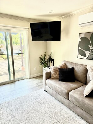 Een woonkamer met een beige bank, een flatscreen televisie aan de muur, een potplant en een groot raam met uitzicht.