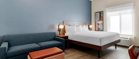 A hotel room with a bed, a sofa, an ottoman, a nightstand, a wall art piece, and a window.