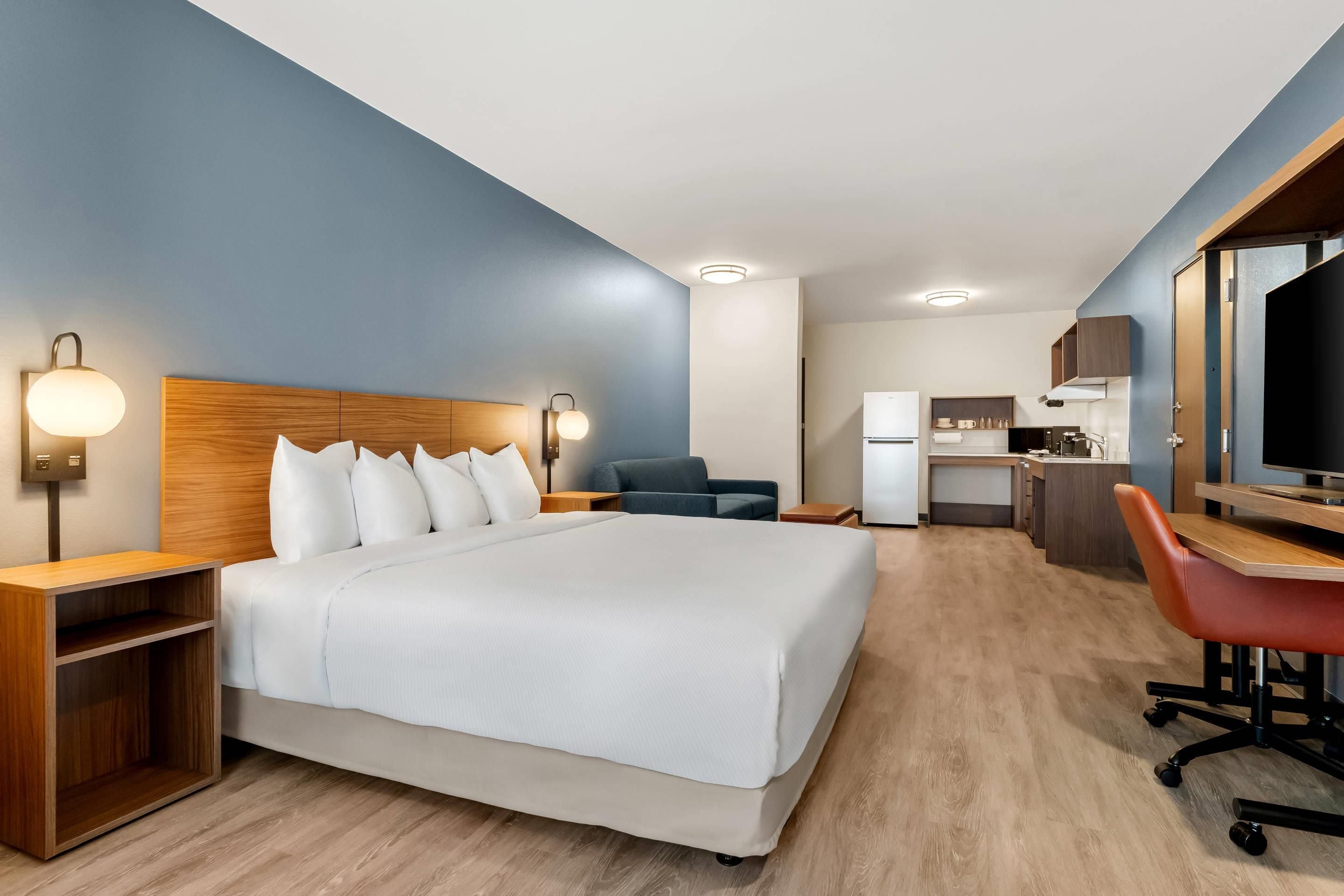 A hotel room with a bed, bedside tables, a desk with a chair, a kitchenette, and a television.