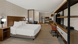 Una habitación de hotel con cama, mesita de noche, lámpara, escritorio, silla y kitchenette.