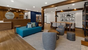 Un moderno vestíbulo de hotel con mostrador de recepción, un sofá azul, sillones grises, un área de comedor y cuadros en la pared.