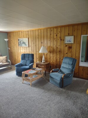 Une pièce aux murs recouverts de lambris en bois, deux fauteuils bleus, une petite table avec des magazines, un lampadaire et un tableau encadré accroché au mur.