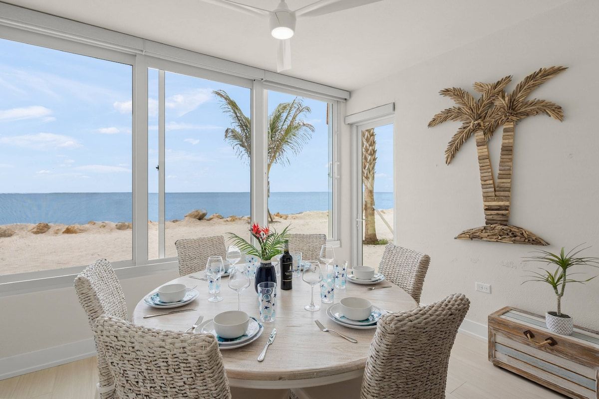 A dining area with a view of the beach, a table set with plates, glasses, and cutlery, and wicker chairs.