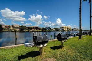 A lakeside view with two barbecue grills on a grassy area, a dock, and buildings in the background. - NEW! Beachfront Ground Floor: Million $ Views (Sarasota)