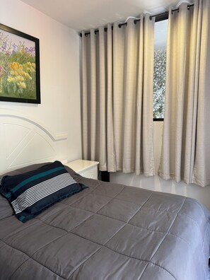 Un dormitorio con cama, mesita de noche, un cuadro y ventana con cortinas.