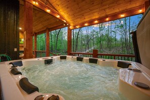 A hot tub with a wooden deck and railings, surrounded by trees. - Moonlit Waves · New! Pond + Deck | Hot Tub, Fire Pit & Cozy Cabin (Broken Bow)