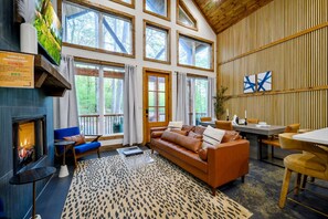 A cozy living room with a fireplace, large windows, a dining area, and a view of the forest. - Moonlit Waves · New! Pond + Deck | Hot Tub, Fire Pit & Cozy Cabin (Broken Bow)