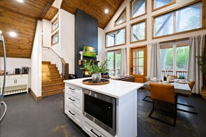 A modern kitchen with a central island, a dining area with a table and chairs, a staircase, and large windows with a view of the outdoors. - Moonlit Waves · New! Pond + Deck | Hot Tub, Fire Pit & Cozy Cabin (Broken Bow)