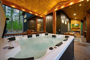 A hot tub in a wooden cabin with a deck, a fireplace, and a TV mounted on the wall. - Moonlit Waves · New! Pond + Deck | Hot Tub, Fire Pit & Cozy Cabin (Broken Bow)