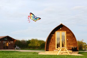 A wooden cabin with a large window and a door, a bicycle parked outside, and a butterfly-shaped kite flying in the sky. - Cosy Rural Cabin with Electric Hot Tub (Grantham)
