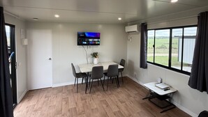 Ein modernes Zimmer mit Esstisch, Stühlen, einem an der Wand befestigten Fernseher, einem Fenster mit Vorhängen und einem kleinen Schreibtisch mit Stuhl.